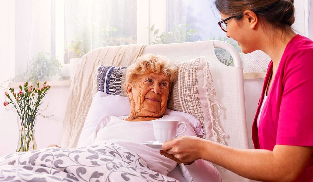 Eine ältere Frau liegt im Bett und lächelt eine Pflegekraft an, die ihr eine Tasse reicht.